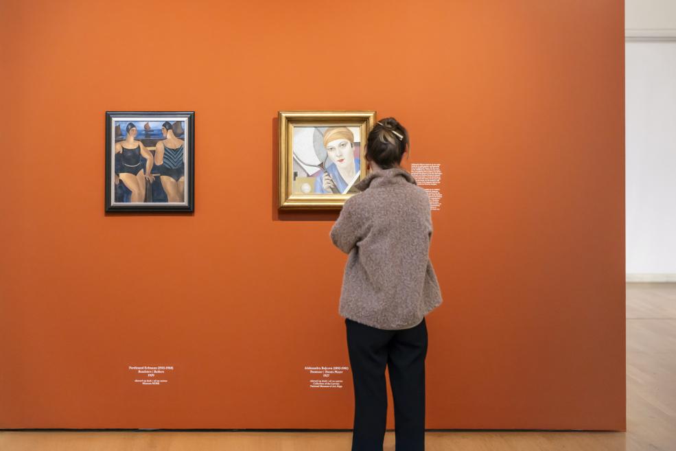 Sieviete muzeja telpā stāv pretī diviem modernisma darbiem, kas eksponēti uz oranžas sienas.