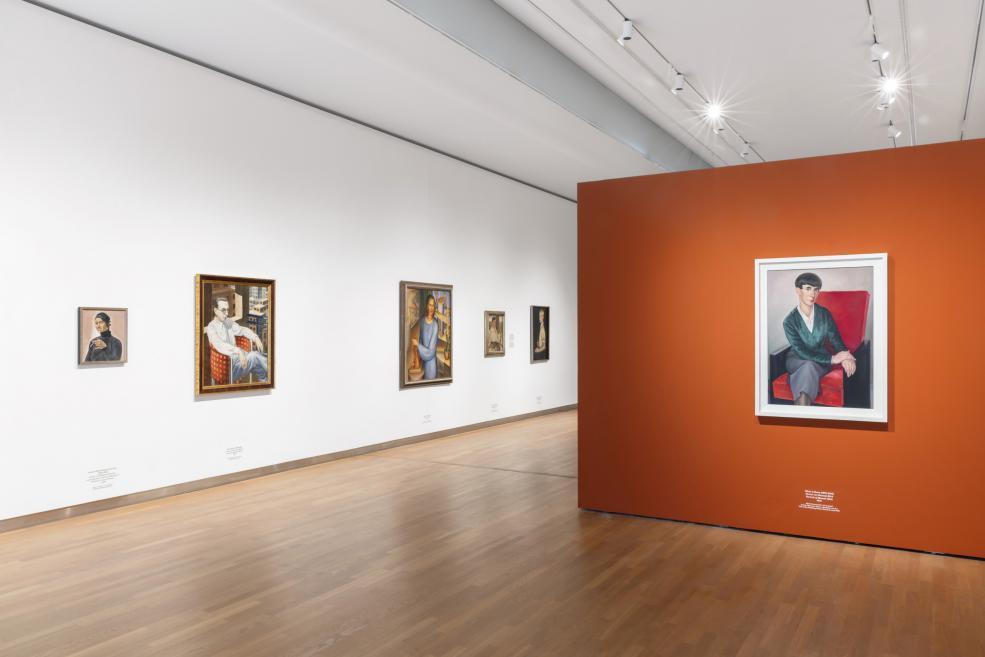 Izstāžu zālē ar modernisma portretiem un centrā novietotu soliņu skatāmas gleznas uz baltas un oranžas sienas.