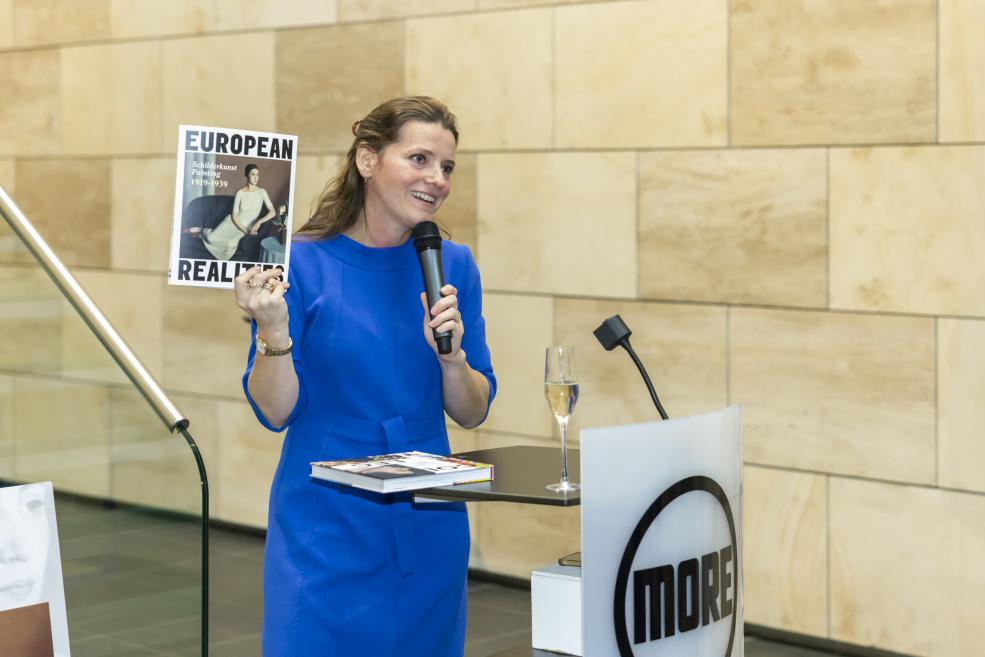 Sieviete zilā kleitā tur mikrofonu un izstādes katalogu, uzstājoties publikas priekšā.