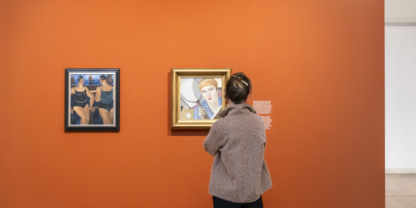 Apmeklētāja stāv pie oranžas sienas un tuvplānā aplūko divus blakus izvietotus gleznu portretus izstāžu zālē.
