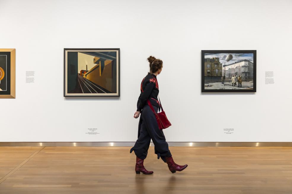 Sieviete pārvietojas gar sienu ar vairākām gleznām plašā, gaišā izstāžu telpā.