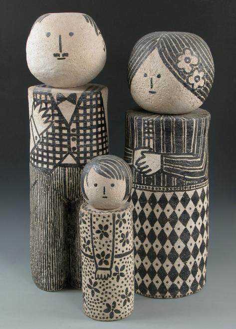 Četras stilizētas koka figūras ar apaļām galvām un melnbaltiem ģeometriskiem un ornamentāliem zīmējumiem uz tumša fona.