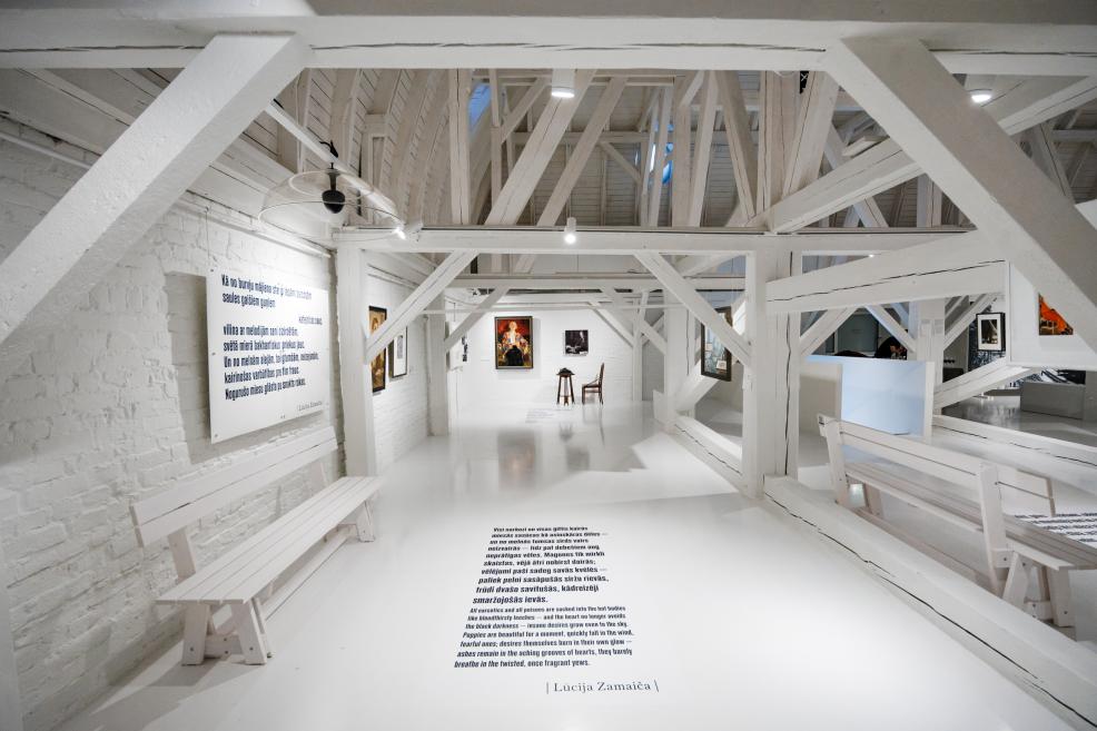 Balta mākslas galerijas ekspozīcijas zāle ar koka sijām, soliņiem, uz sienām izvietotiem darbiem un grīdā iespiestu tekstu.