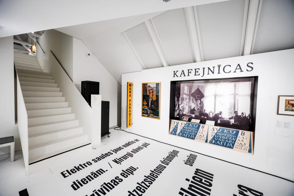 Muzeja izstādes telpa ar baltām kāpnēm, uz sienas uzrakstu KAFEJNĪCAS, lielu melnbaltu fotogrāfiju un vairākiem mākslas darbiem.