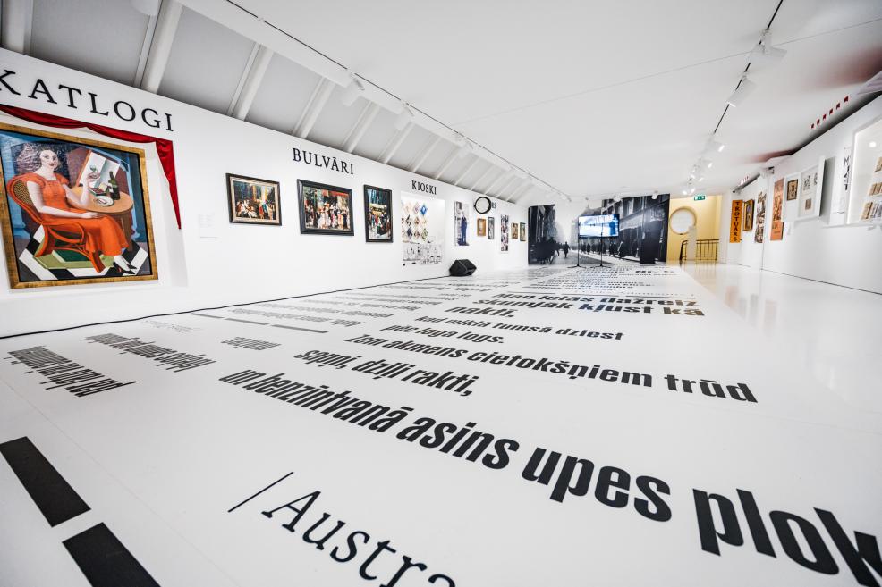 Plaša muzeja izstāžu zāle ar pie sienām izvietotām gleznām, plakātiem un melnbaltu foto instalāciju, bet uz baltās grīdas redzams liels drukāts latviešu teksts.