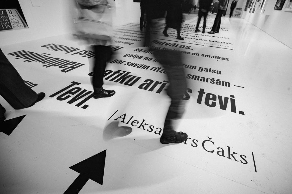 Melnbalts muzeja interjera skats ar staigājošiem apmeklētājiem un uz grīdas un sienām izvietotu lielu tipogrāfisku tekstu.