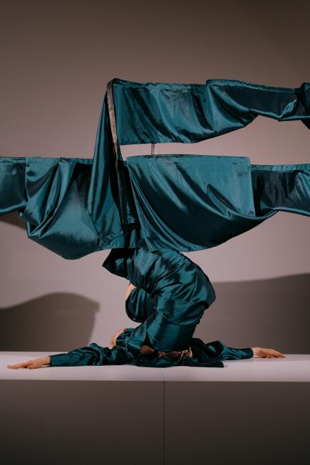 Skatuves performance ar figūru spoži zaļganzilā satīna tērpā, kas saliekts abstraktā pozā uz balta paaugstinājuma pret pelēku fonu.