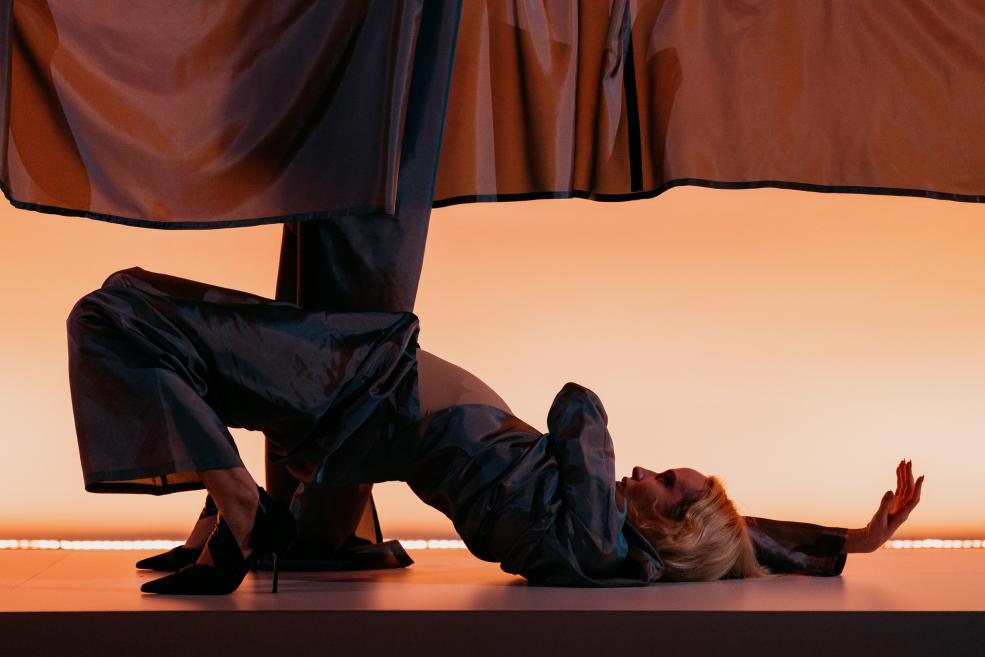 Performances mākslas instalācijā redzama blondmataina sieviete melnā tērpā guļus pozā uz paaugstinājuma zem piekārta tumša auduma, uz oranža fona.