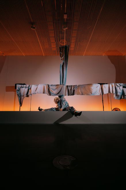 Skatuves performance ar blondmatainu izpildītāju spīdīgā zilā tērpā, sēžot uz paaugstinājuma zem piekārtām sudrabainām auduma joslām un koka līstēm izgaismotā telpā.