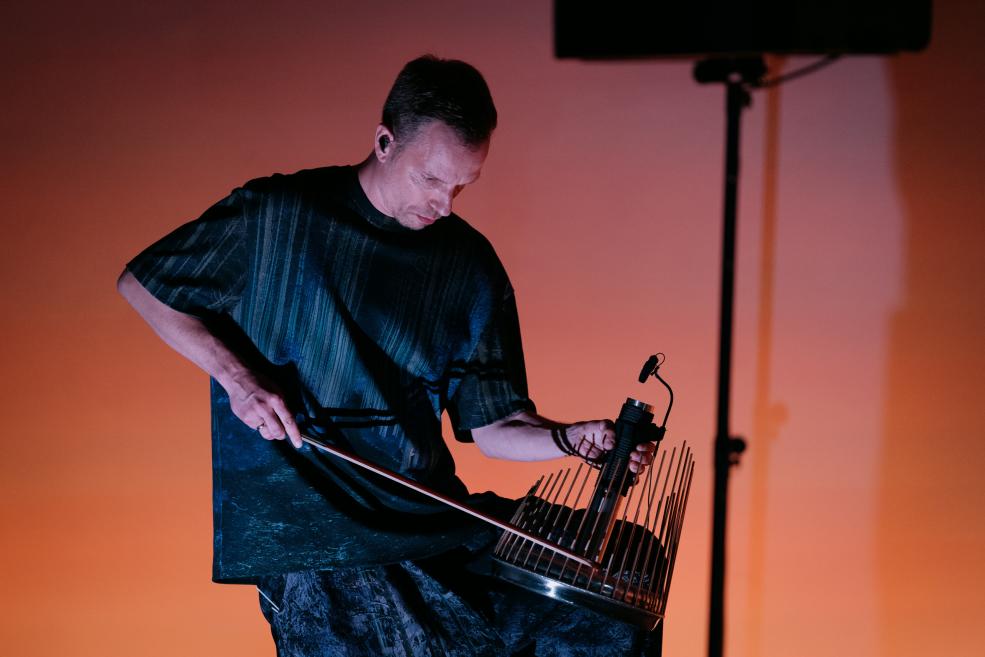 Vīrietis uz skatuves spēlē eksperimentālu stīgu instrumentu oranžā apgaismojumā.
