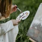 Painting supplies with brushes, paint tubes and a small landscape painting on a canvas on an easel outdoors.