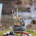 A person in a cap paints a landscape in watercolor outdoors, using a paint set and paper on a cardboard support.