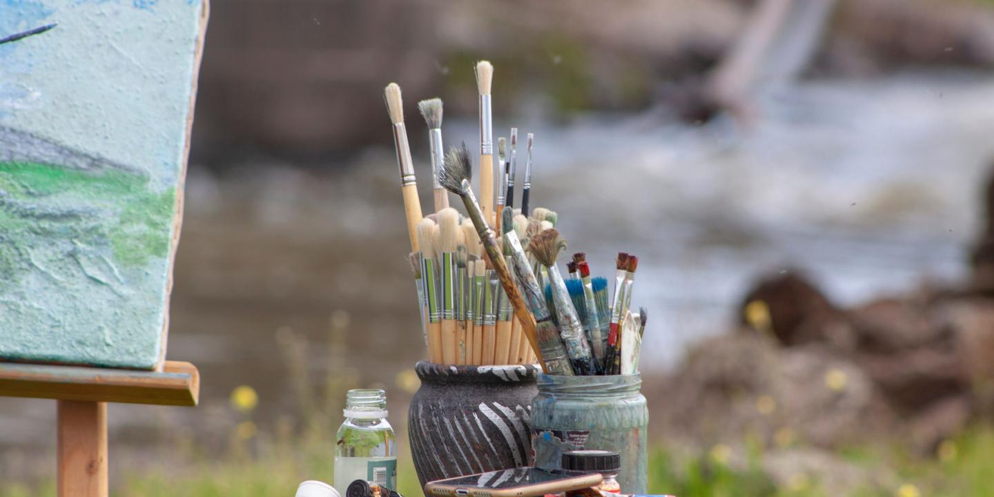 A person in a cap paints a landscape in watercolor outdoors, using a paint set and paper on a cardboard support.