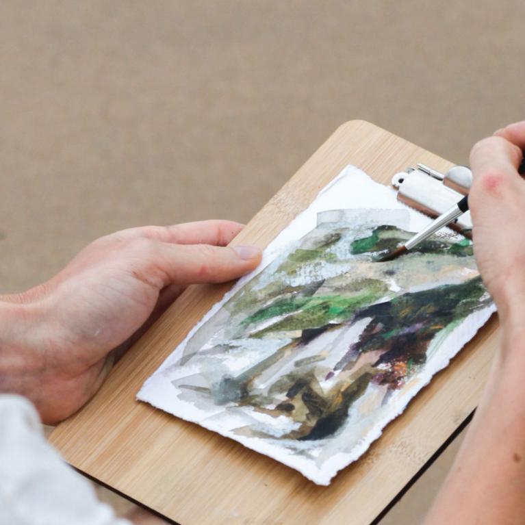 Hands paint on a watercolor sketch pinned to a wooden board with a brush.