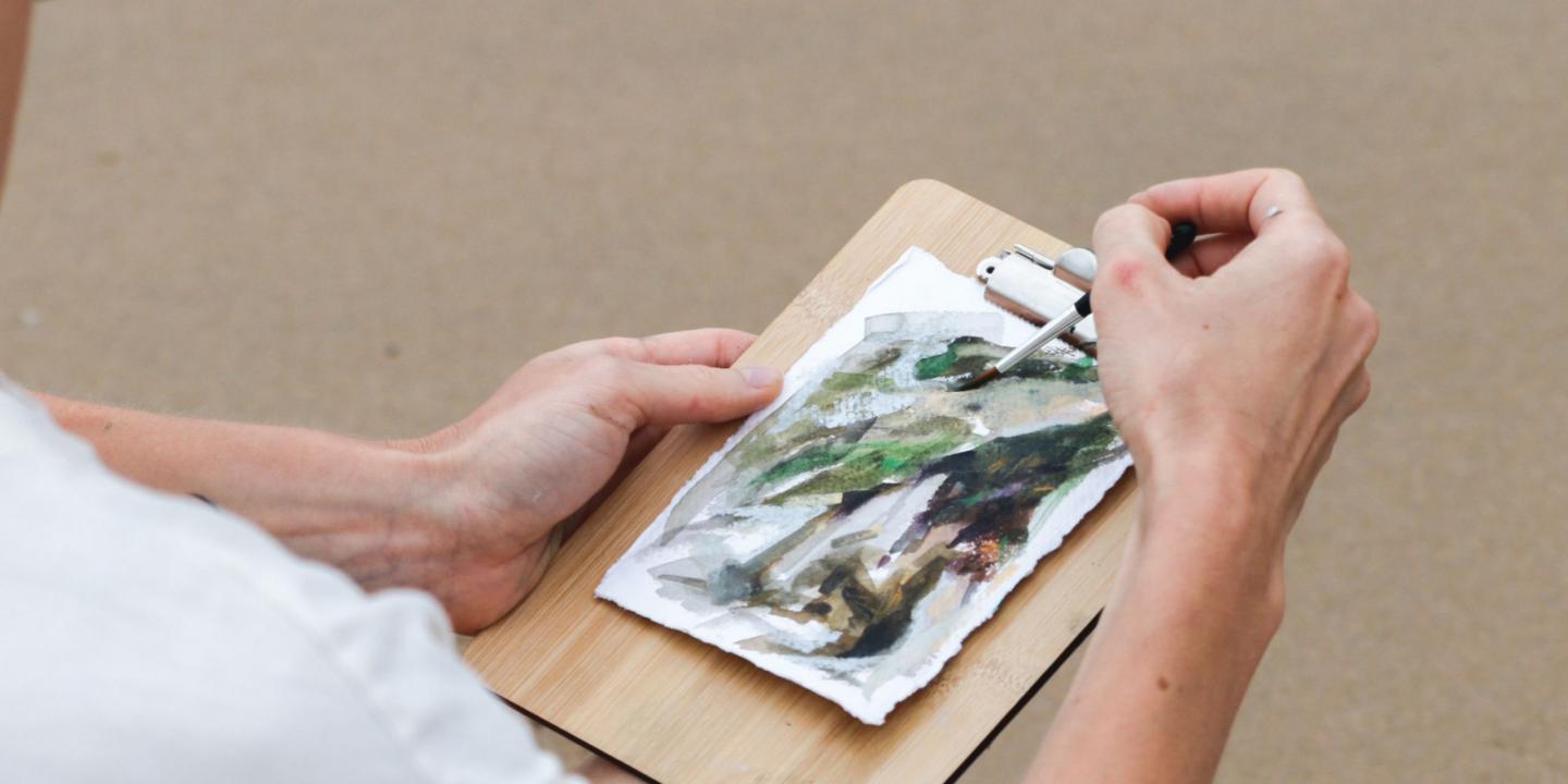 Hands paint on a watercolor sketch pinned to a wooden board with a brush.