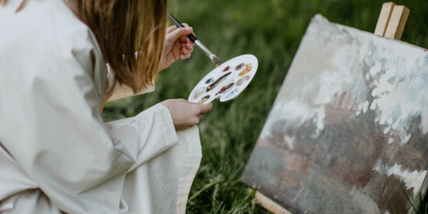Painting supplies with brushes, paint tubes and a small landscape painting on a canvas on an easel outdoors.