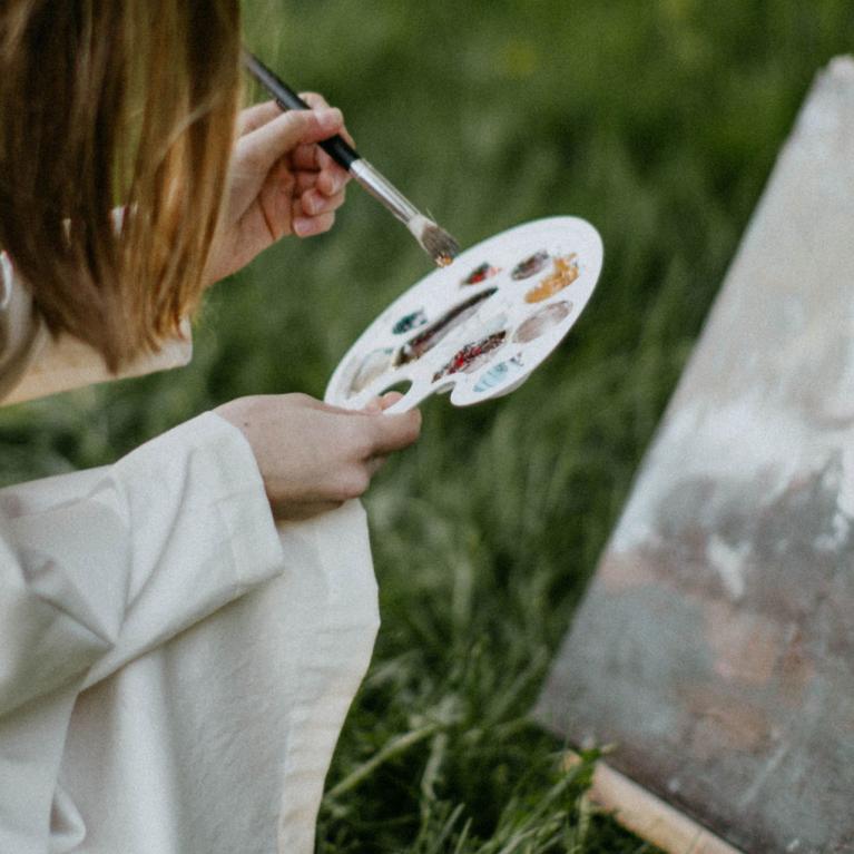 Painting supplies with brushes, paint tubes and a small landscape painting on a canvas on an easel outdoors.