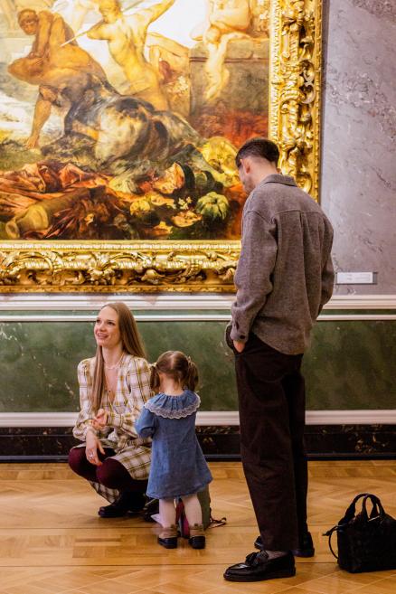 Sieviete ar bērnu un vīrietis stāv pie gleznas muzejā.