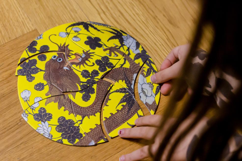 Dzeltena apaļas formas puzle ar brūna pūķa attēlu uz koka grīdas.