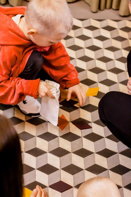 Zēns sarkana džempera krāsā saliek krāsainas figūras uz grīdas.