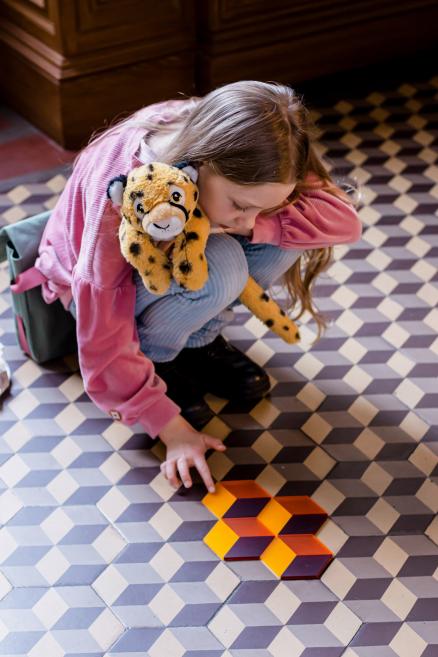 Meitene ar plīša gepardu veido figūru no krāsainiem laukumiem uz flīžu grīdas.