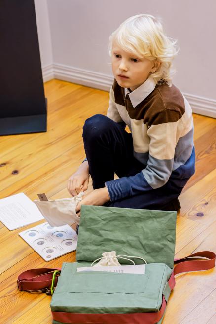 Zēns ar zaļu muzeja somu pēta uzdevumu lapas un maisiņu.