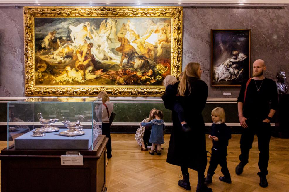 Ģimene ar bērniem aplūko gleznu zeltītā rāmī muzejā.