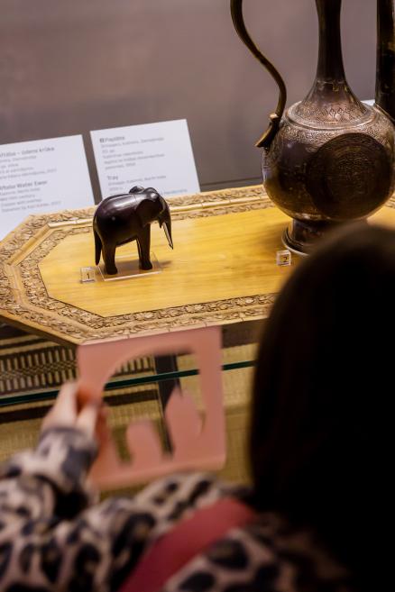Meitene ar rozā rāmīti skatās uz ziloņa figūru vitrīnā.