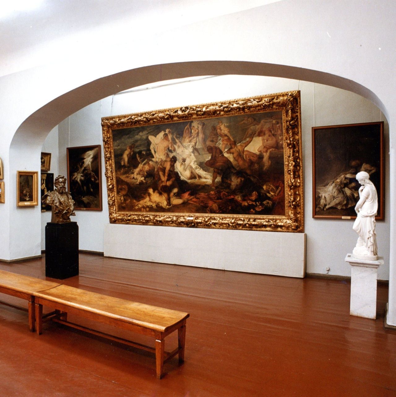 Museum gallery with a monumental historical painting in an ornate frame, sculptures, and a bench for visitors.