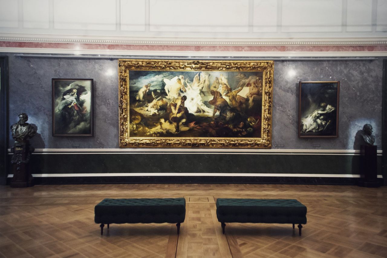 Museum gallery interior with a monumental historical painting in an ornate frame, two smaller paintings, and sculptural busts.