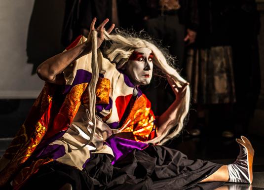 A stage performer in a vivid costume and makeup strikes a dramatic pose during a performance while holding long white hair.