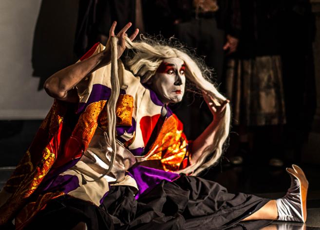 A stage performer in a vivid costume and makeup strikes a dramatic pose during a performance while holding long white hair.