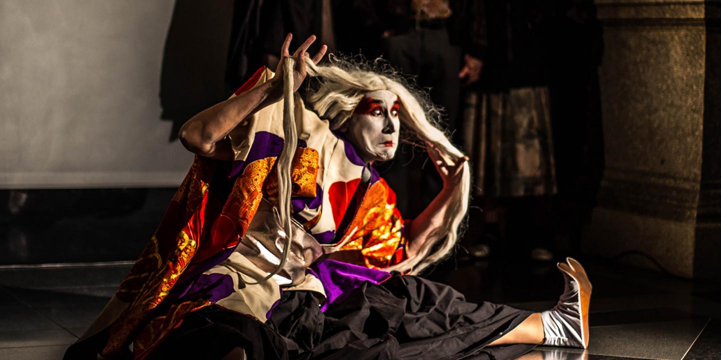 A stage performer in a vivid costume and makeup strikes a dramatic pose during a performance while holding long white hair.