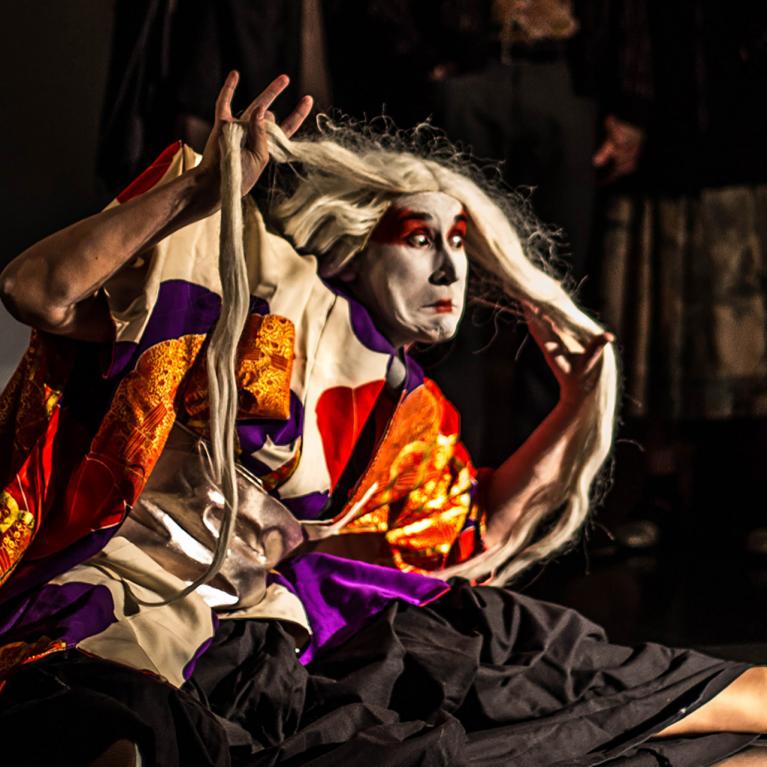 A stage performer in a vivid costume and makeup strikes a dramatic pose during a performance while holding long white hair.