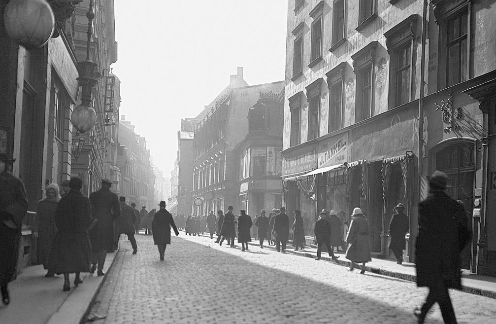 A black-and-white photograph of a busy Riga street in the 1920s: pedestrians in dark coats and hats walk along a cobblestone road, with shop signs and multi-storey buildings lining both sides, softened by morning haze in the distance.