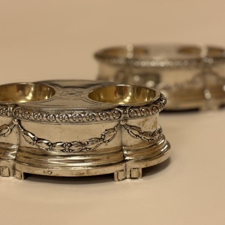 Sudraba trauks ar diviem apaļiem iedobumiem, rotāts ar reljefu ornamentu, uz neitrāla fona; fonā redzams otrs līdzīgs trauks.