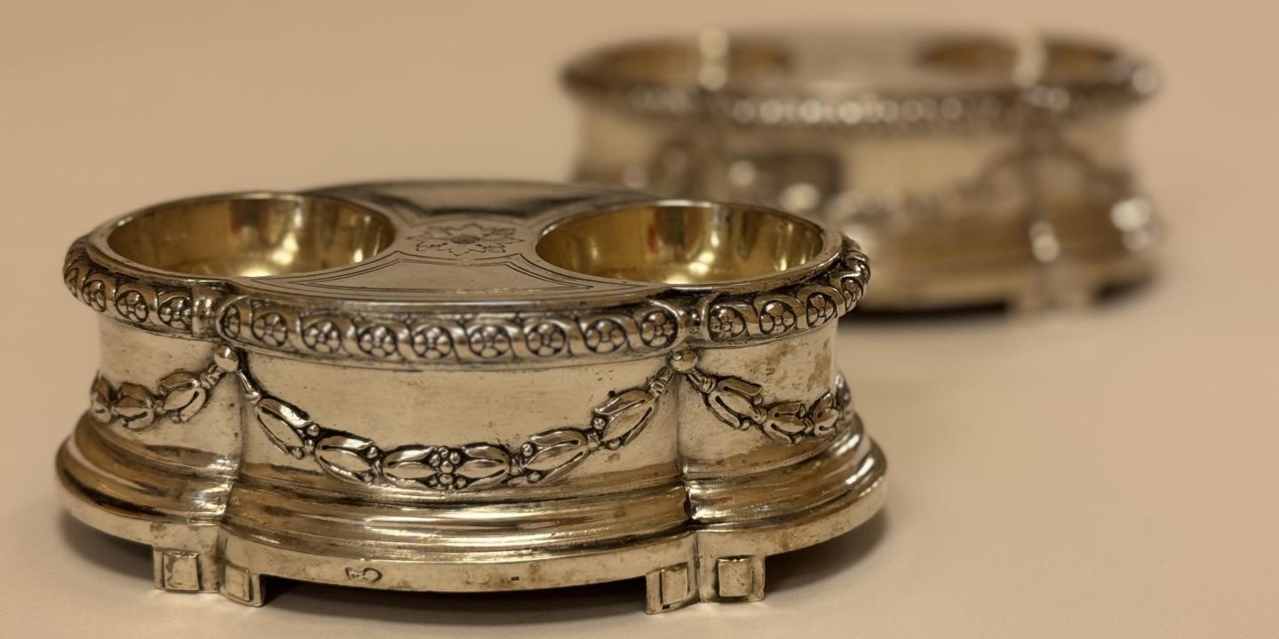 Sudraba trauks ar diviem apaļiem iedobumiem, rotāts ar reljefu ornamentu, uz neitrāla fona; fonā redzams otrs līdzīgs trauks.