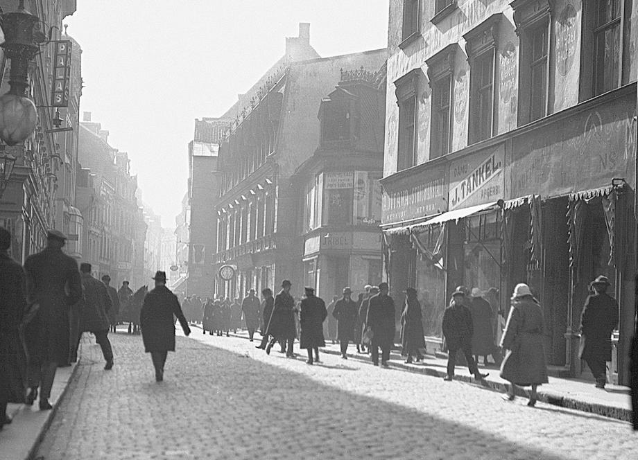 A black-and-white photograph of a busy Riga street in the 1920s: pedestrians in dark coats and hats walk along a cobblestone road, with shop signs and multi-storey buildings lining both sides, softened by morning haze in the distance.