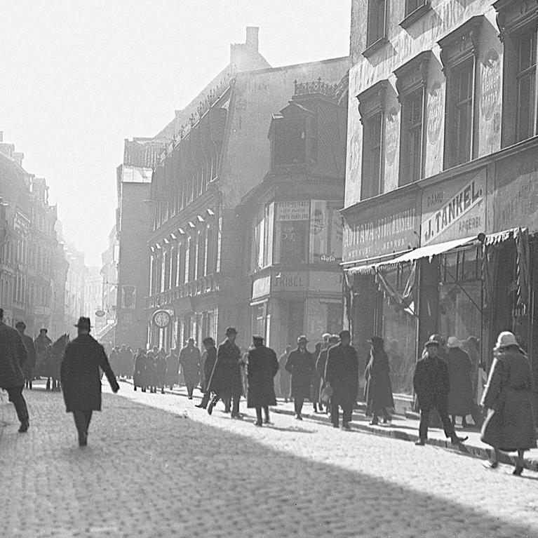 A black-and-white photograph of a busy Riga street in the 1920s: pedestrians in dark coats and hats walk along a cobblestone road, with shop signs and multi-storey buildings lining both sides, softened by morning haze in the distance.