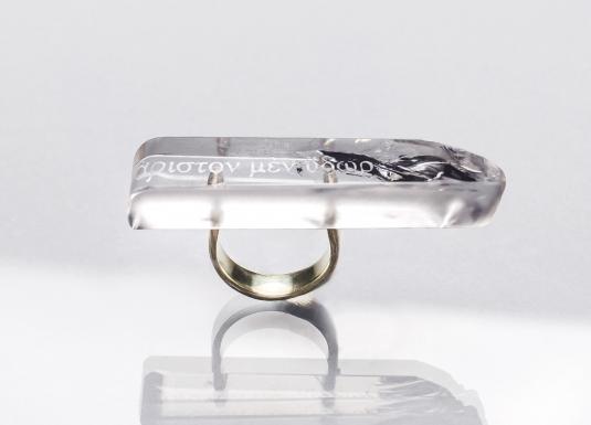 Caurspīdīga taisnstūrveida gredzena galviņa ar iestrādātu tekstu un tumšu dekoratīvu elementu, piestiprināta pie metāla gredzena uz gaiša fona ar atspulgu.