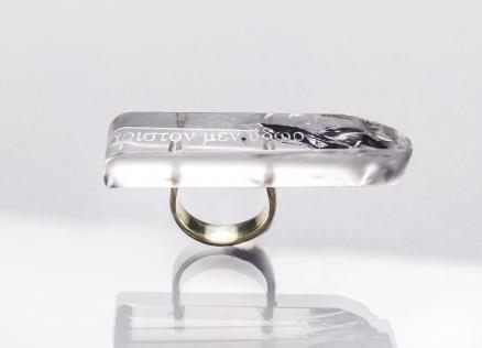 Caurspīdīga taisnstūrveida gredzena galviņa ar iestrādātu tekstu un tumšu dekoratīvu elementu, piestiprināta pie metāla gredzena uz gaiša fona ar atspulgu.