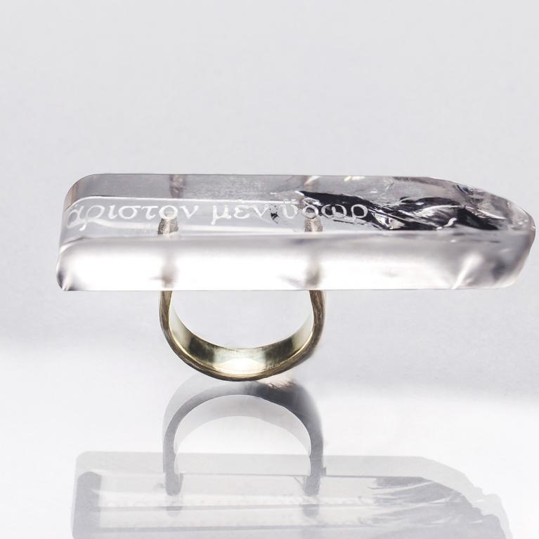 Caurspīdīga taisnstūrveida gredzena galviņa ar iestrādātu tekstu un tumšu dekoratīvu elementu, piestiprināta pie metāla gredzena uz gaiša fona ar atspulgu.