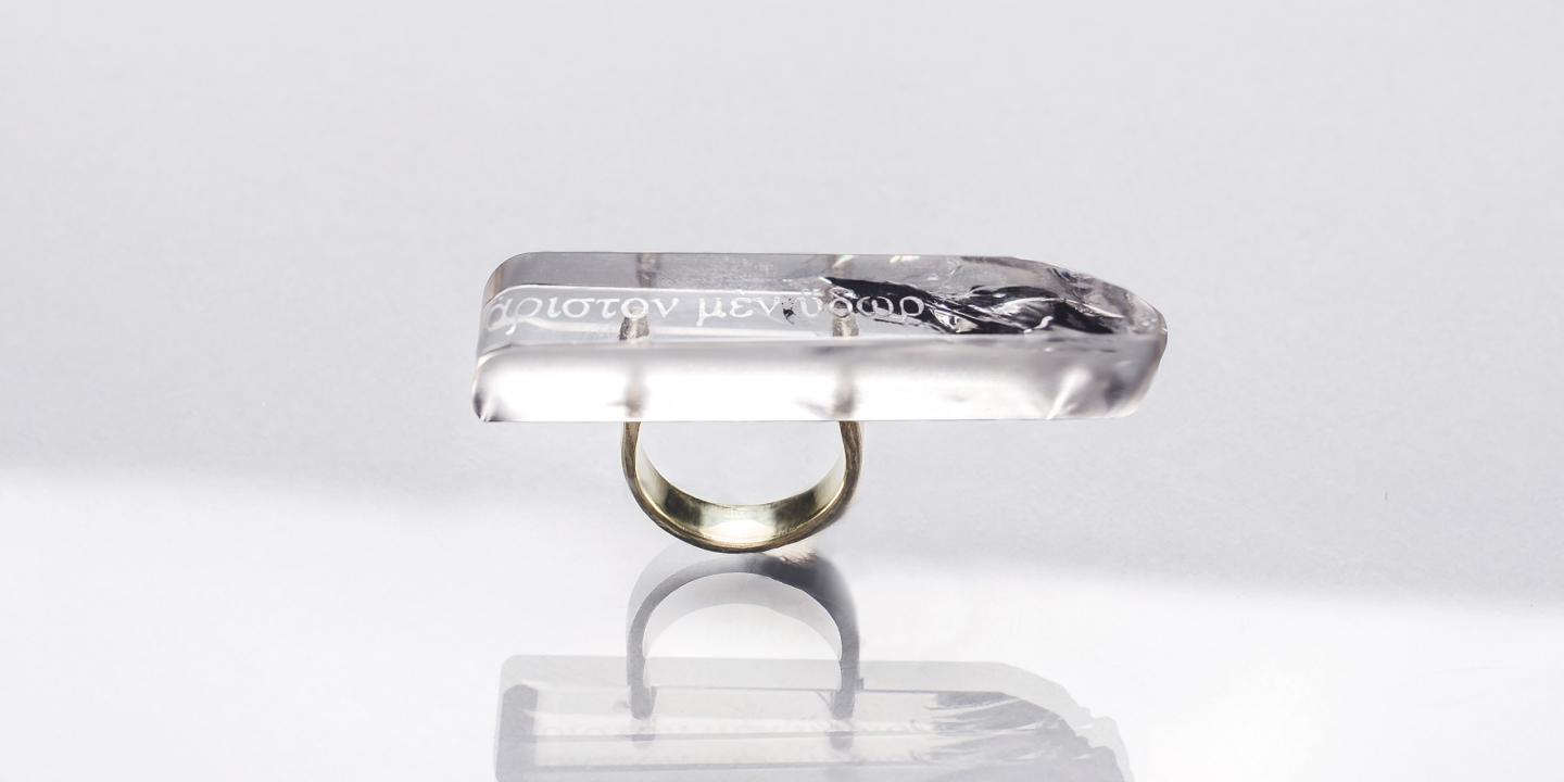 Caurspīdīga taisnstūrveida gredzena galviņa ar iestrādātu tekstu un tumšu dekoratīvu elementu, piestiprināta pie metāla gredzena uz gaiša fona ar atspulgu.