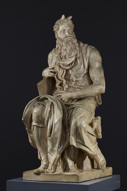 A seated bearded male sculpture with long hair and horns, shown on a dark background on a base.