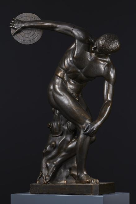 Dark bronze sculpture of a nude male figure leaning forward and holding a round disk with one outstretched arm against a dark background.
