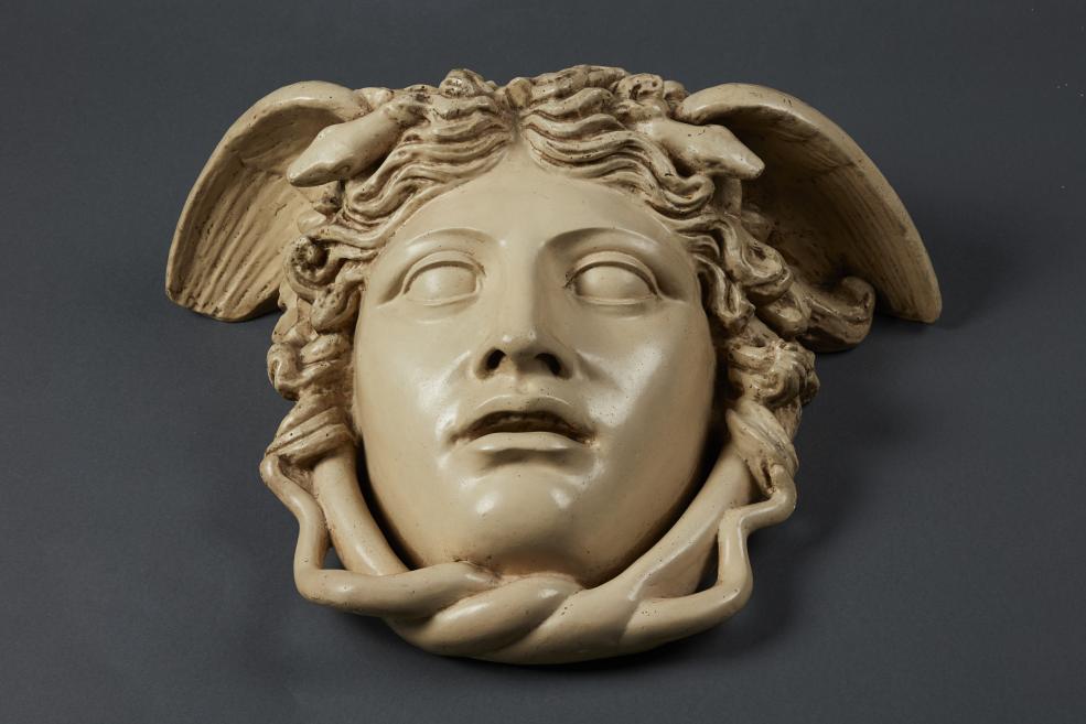 Ceramic mask with a woman’s face, wavy hair, and wings on a dark background.