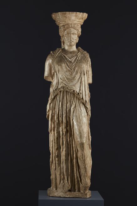 Ancient terracotta statue of a woman with a dish on her head, dressed in a long draped chiton, against a dark background.