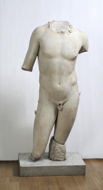 White male torso without head and both arms, a sculpture on a plinth against a white wall.
