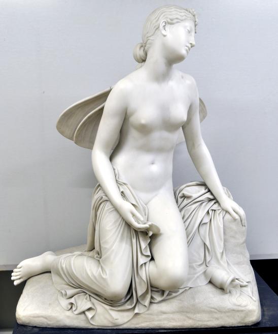 White marble sculpture depicting a seated nude female figure with wings on a stone base.