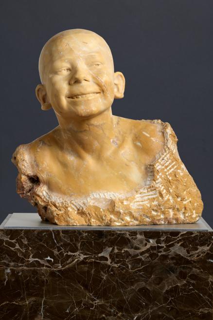 Carved bust of a smiling child’s head and torso in yellowish stone, displayed on a dark marble pedestal against a gray background.